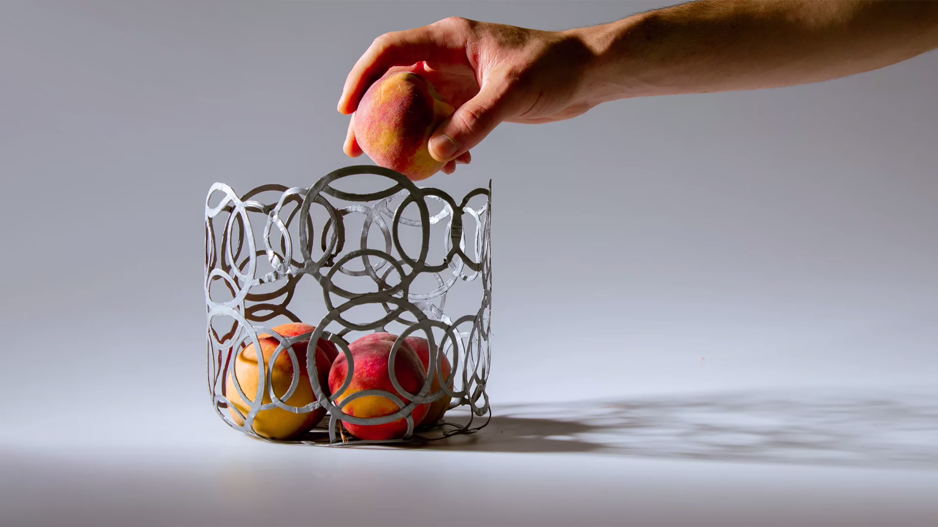 A hand dropping a peach into the PLArt fruit bowl, a shell of fused silver rings